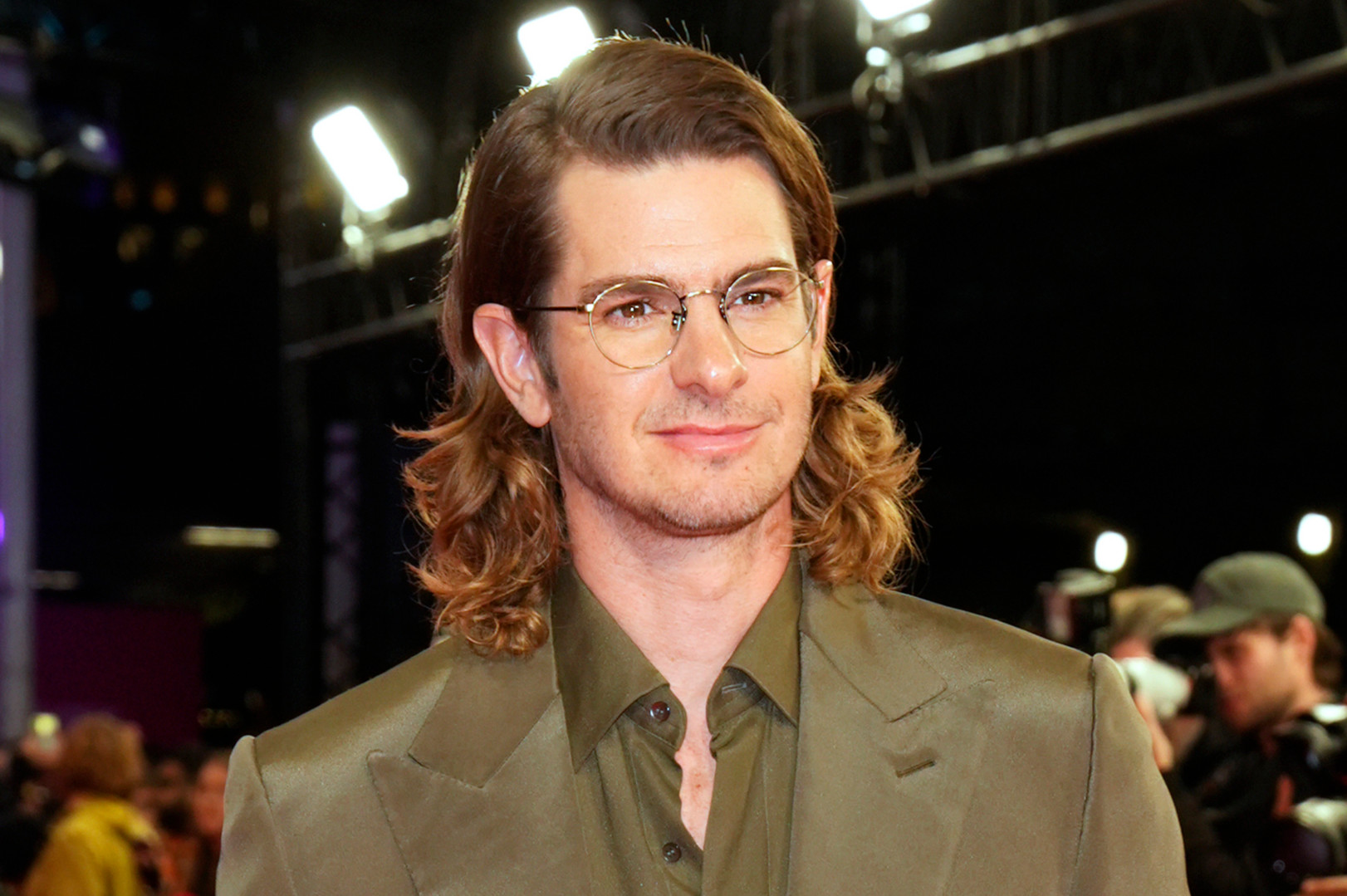Andrew Garfield with glasses and long hair wearing a green suit, standing at an event with bright lights and people in the background.