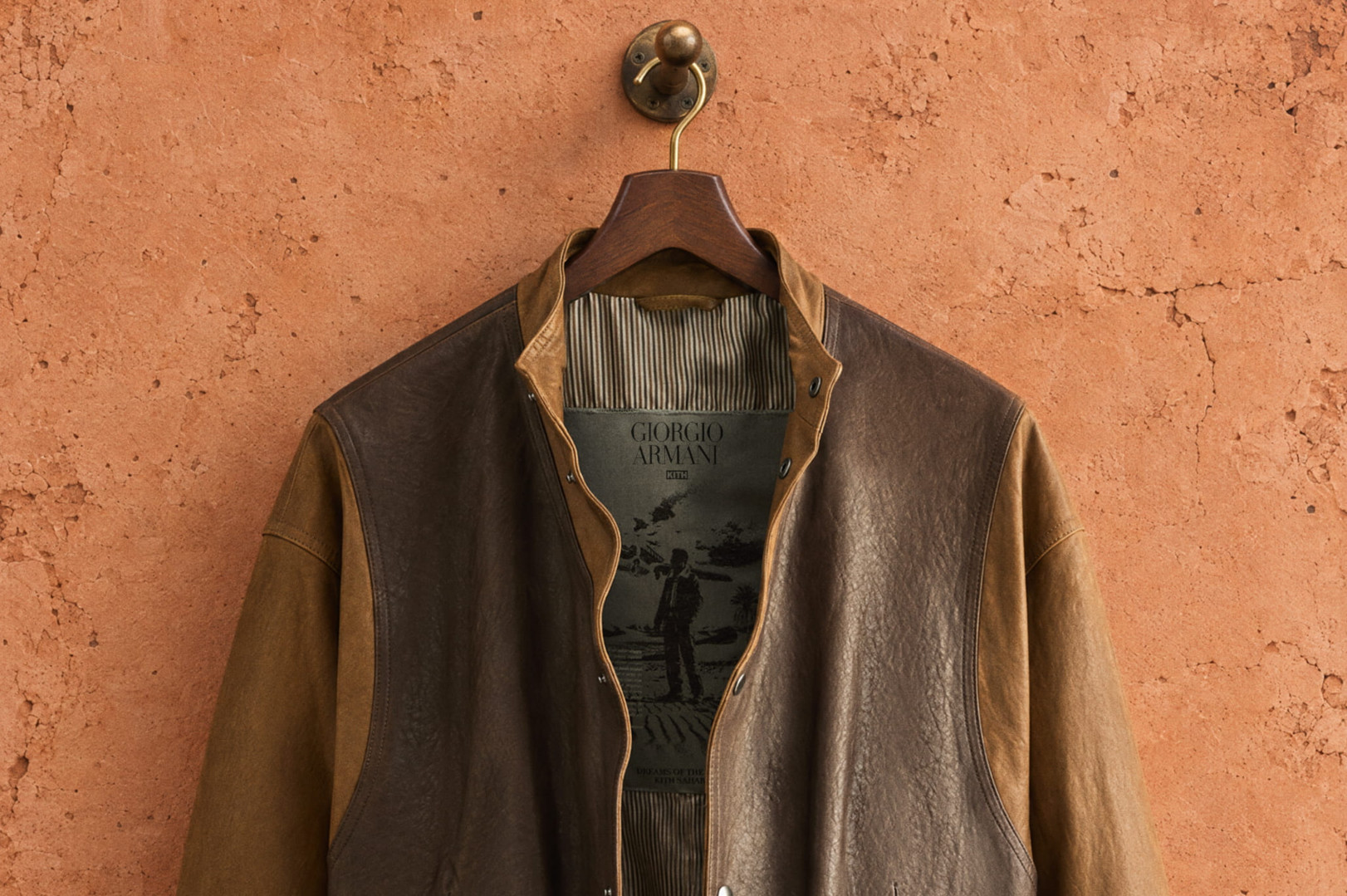 A brown leather Giorgio Armani jacket hangs on a wooden hanger against a textured, rustic wall.