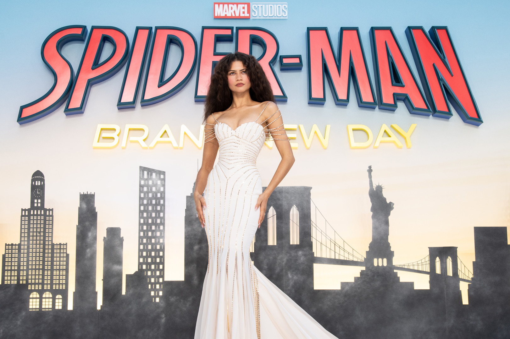 Zendaya in a stunning white gown stands in front of a "Spider-Man: Brand New Day" backdrop featuring a city skyline.