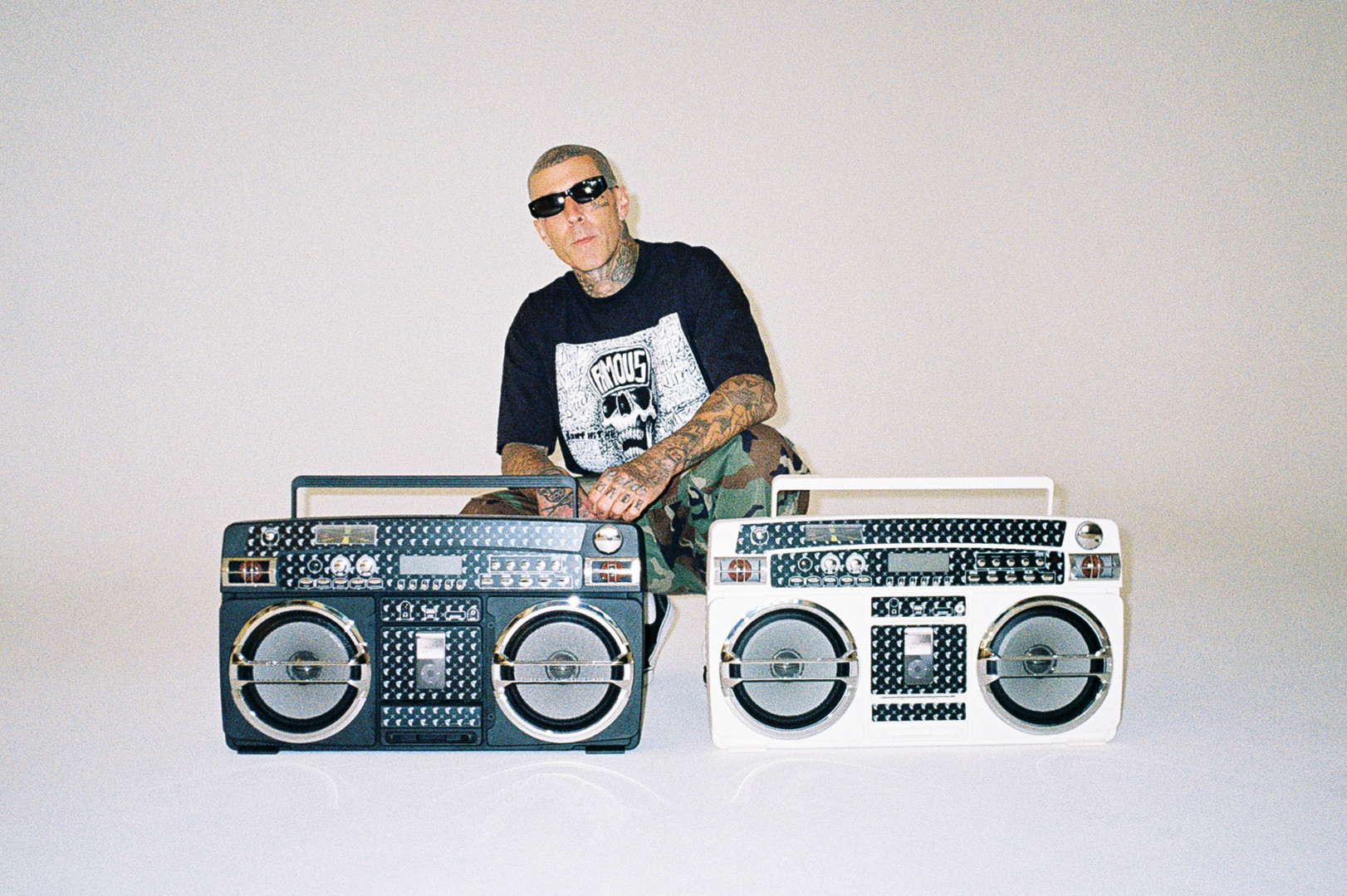 Travis Barker with tattoos and sunglasses sits behind two retro boomboxes, one black and one white, against a plain background.