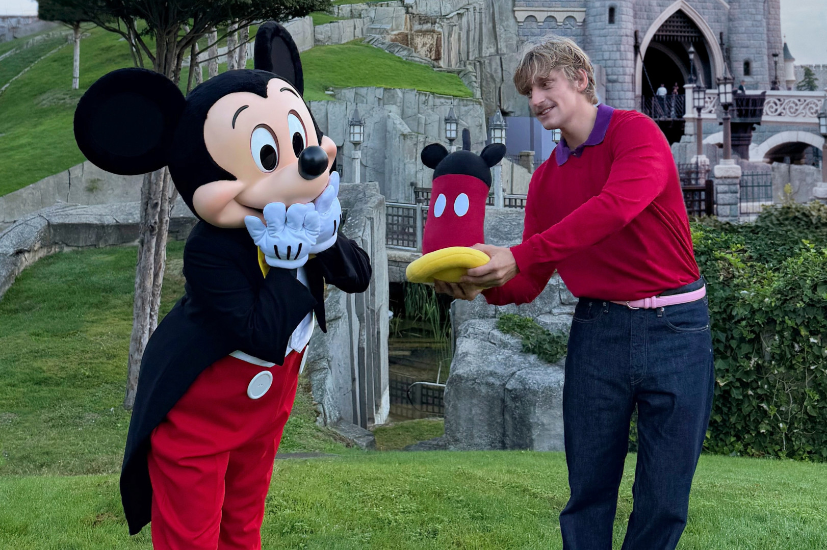 A person in a red sweater hands a Mickey Mouse hat to Mickey Mouse at a theme park, with a castle in the background.