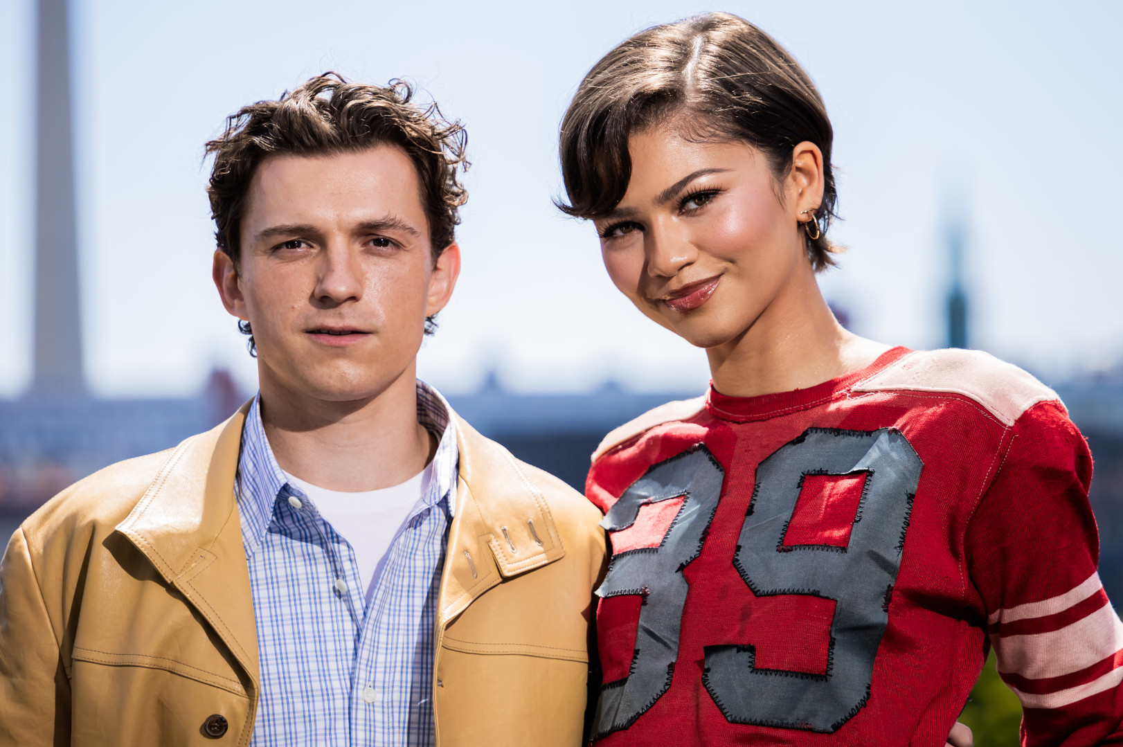 Tom Holland and Zendaya pose outdoors. Tom wears a tan jacket, Zendaya a red top with "39". Both smile against a cityscape backdrop.