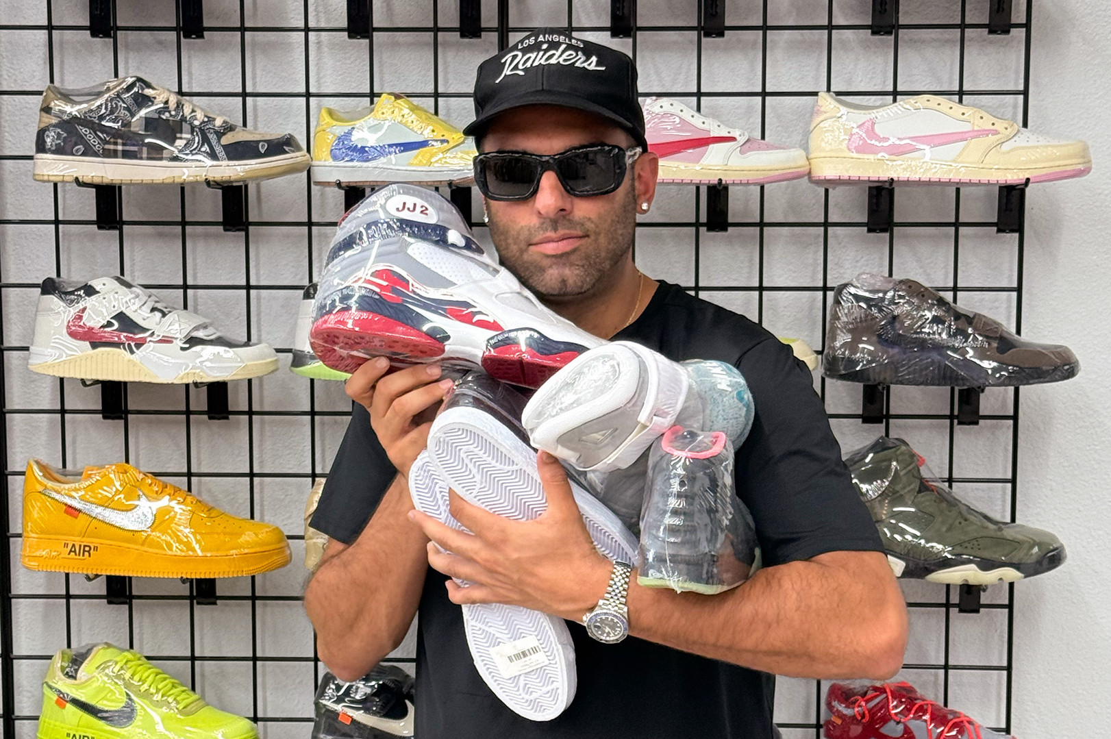 A man wearing sunglasses and a Raiders cap holds several sneakers, standing in front of a wall display of various colorful shoes.