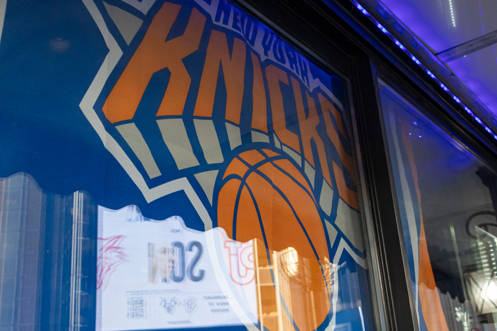 New York Knicks logo on a window, featuring a large basketball and bold orange letters. Reflections and lighting create a dynamic effect.