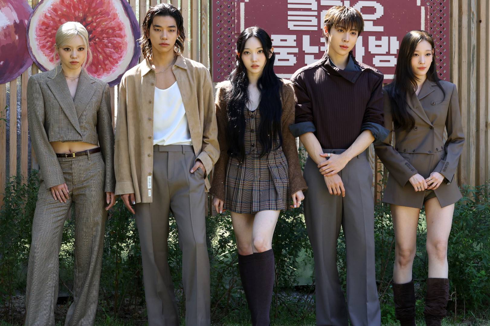ALLDAY PROJECT members stand outdoors in stylish outfits, posing confidently. A wooden fence and large fruit images are in the background.