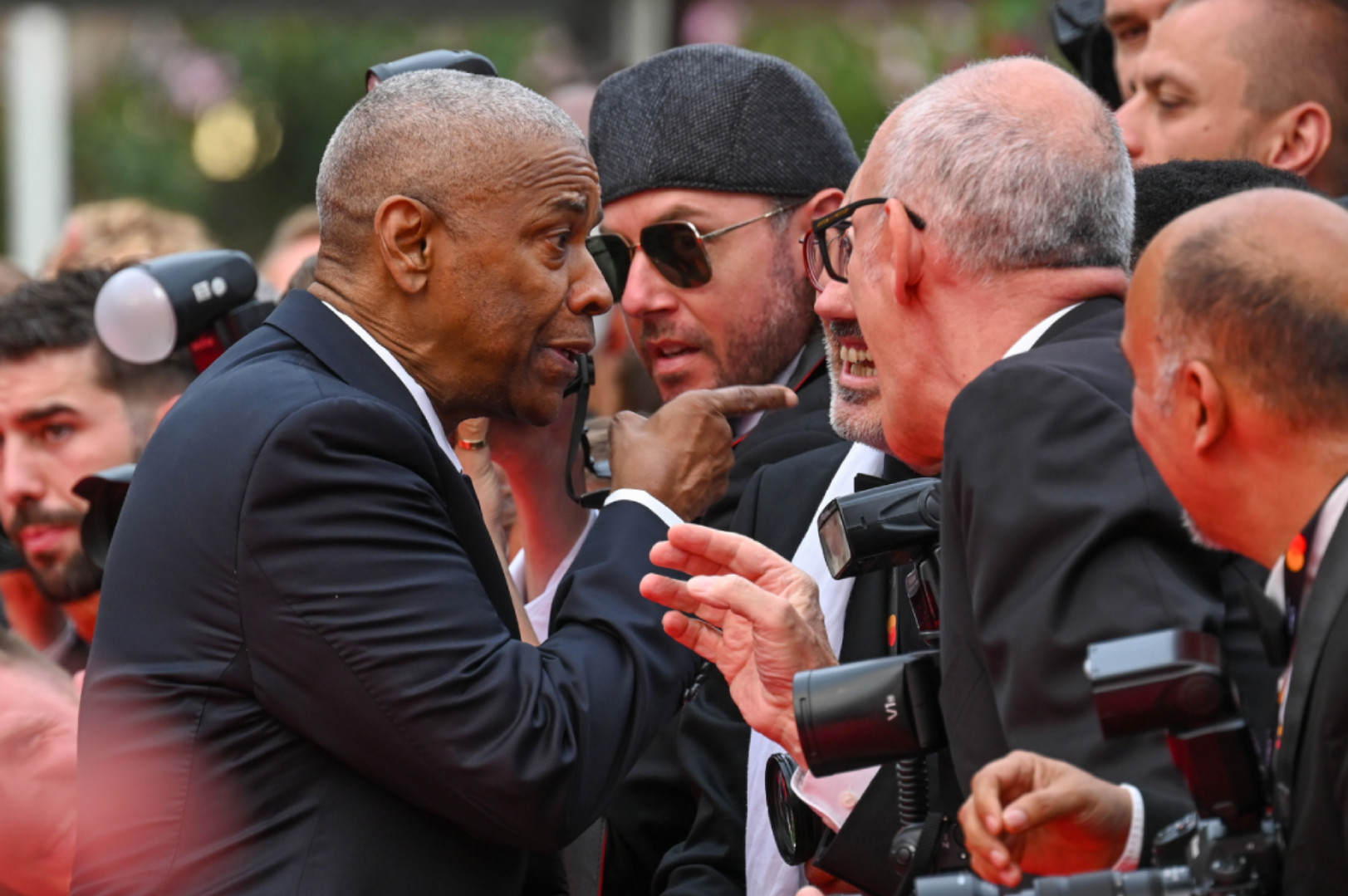 A group of men in suits and ties are engaged in a lively conversation, surrounded by photographers. The scene appears to be at a formal event.