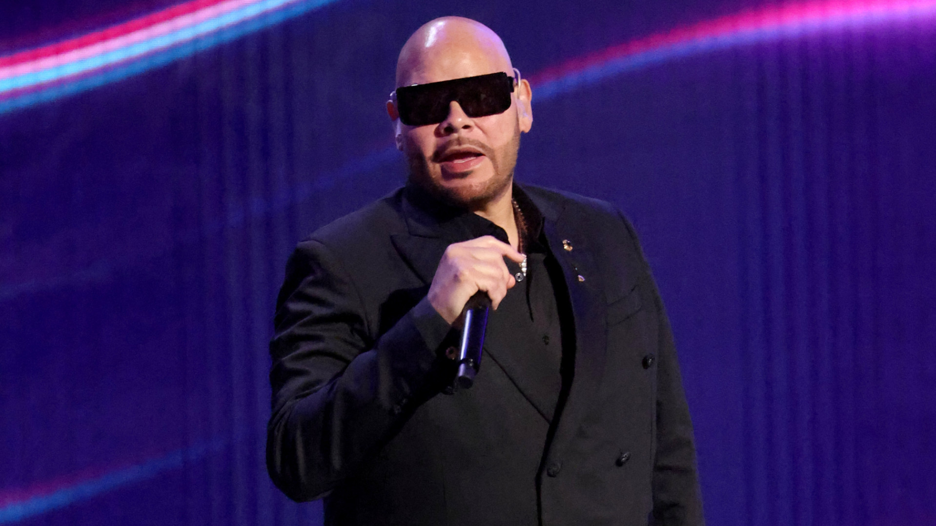 Fat Joe, wearing a black suit and sunglasses, holds a microphone on stage with a colorful background.
