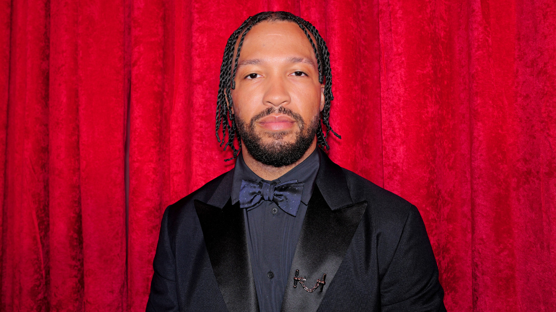 Jalen Brunson with braided hair and a beard in a black suit and bow tie stands against a red curtain backdrop.