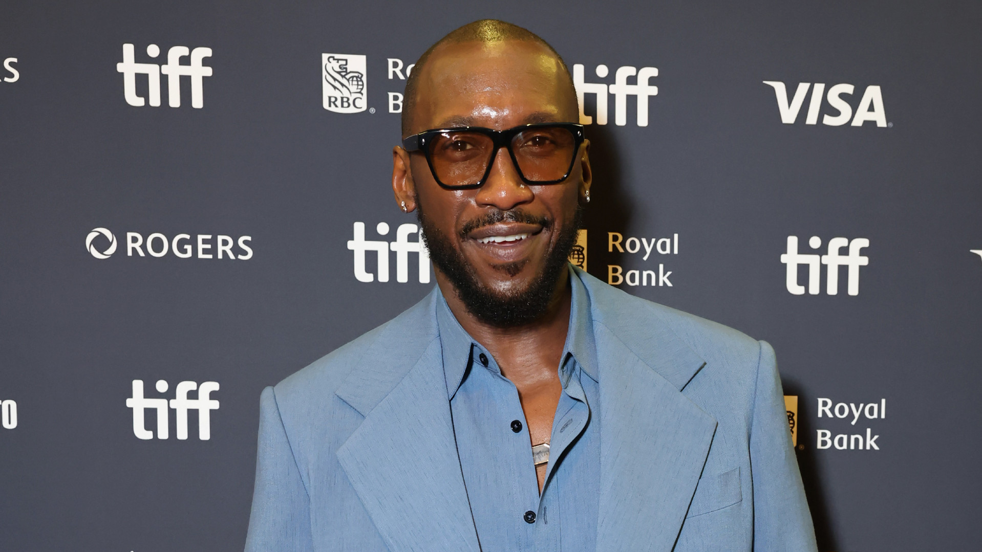 Mahershala Ali in a light blue suit, smiling at the TIFF event backdrop.