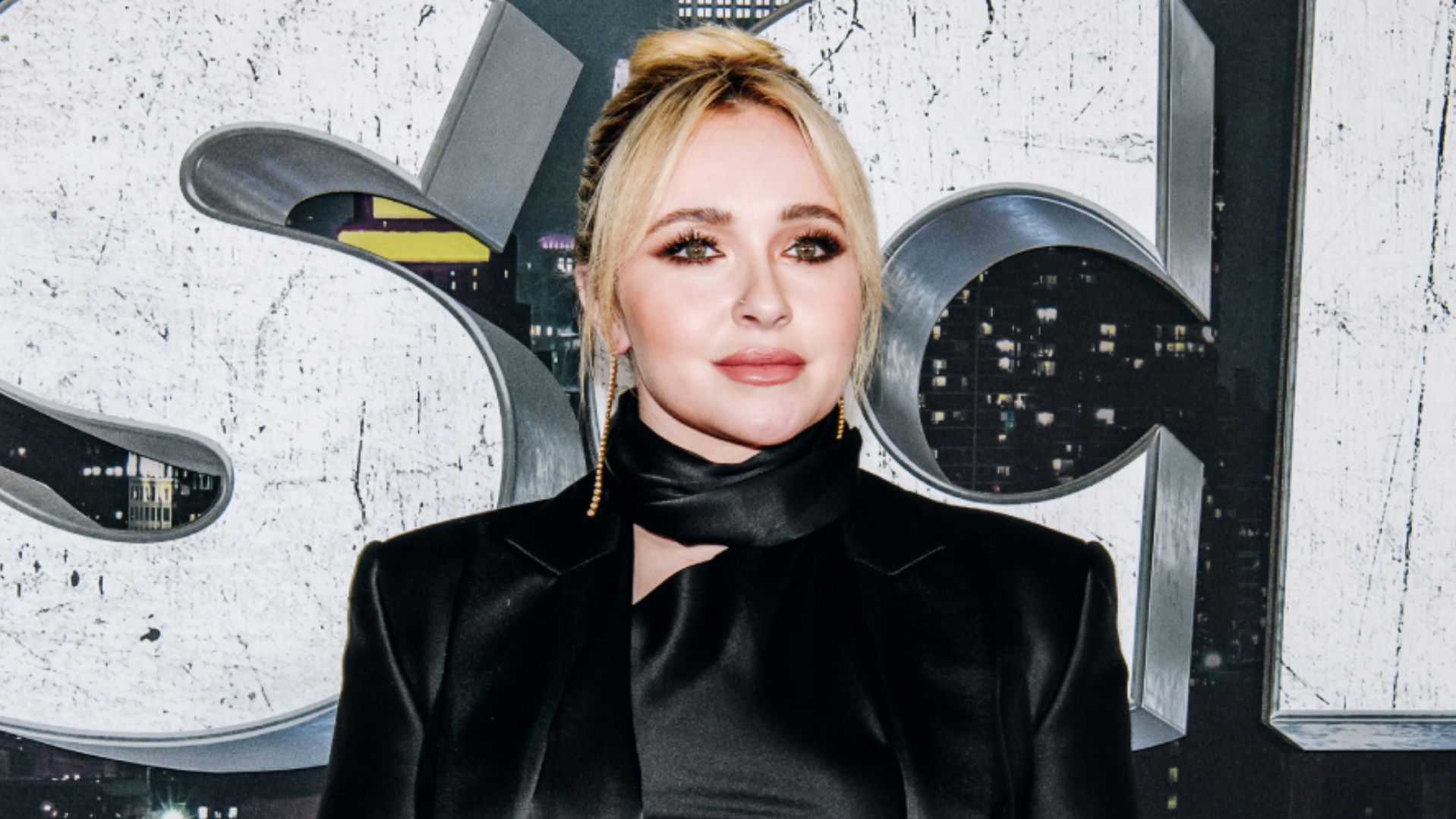 Hayden Panettiere in a black outfit stands in front of a textured backdrop with large letters and city lights.