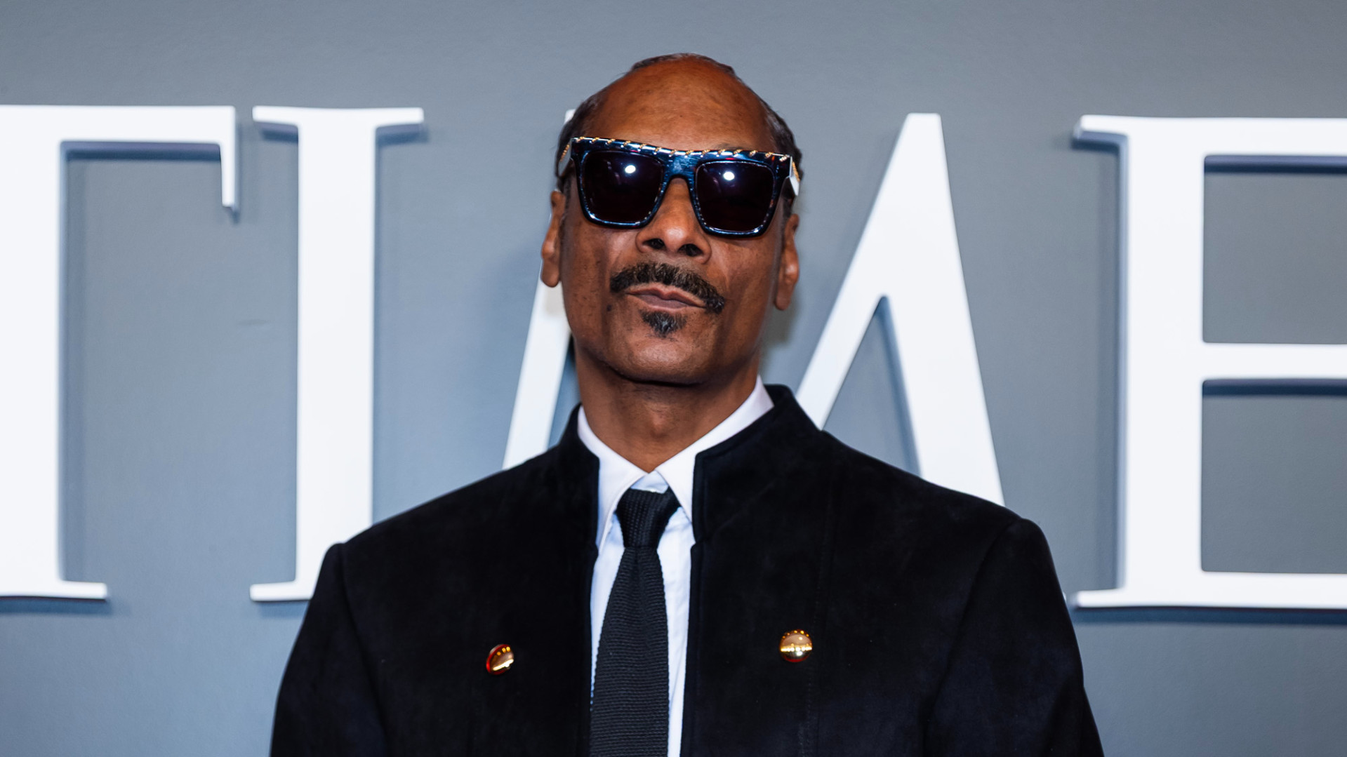 Snoop Dogg wearing sunglasses and a black suit, standing in front of a gray background with white lettering.