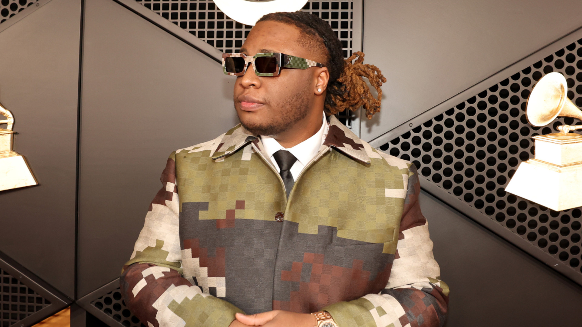 Tay Keith, circa 2024, in a camouflage-patterned coat and sunglasses standing in front of the Grammy Award's step-and-repeat.