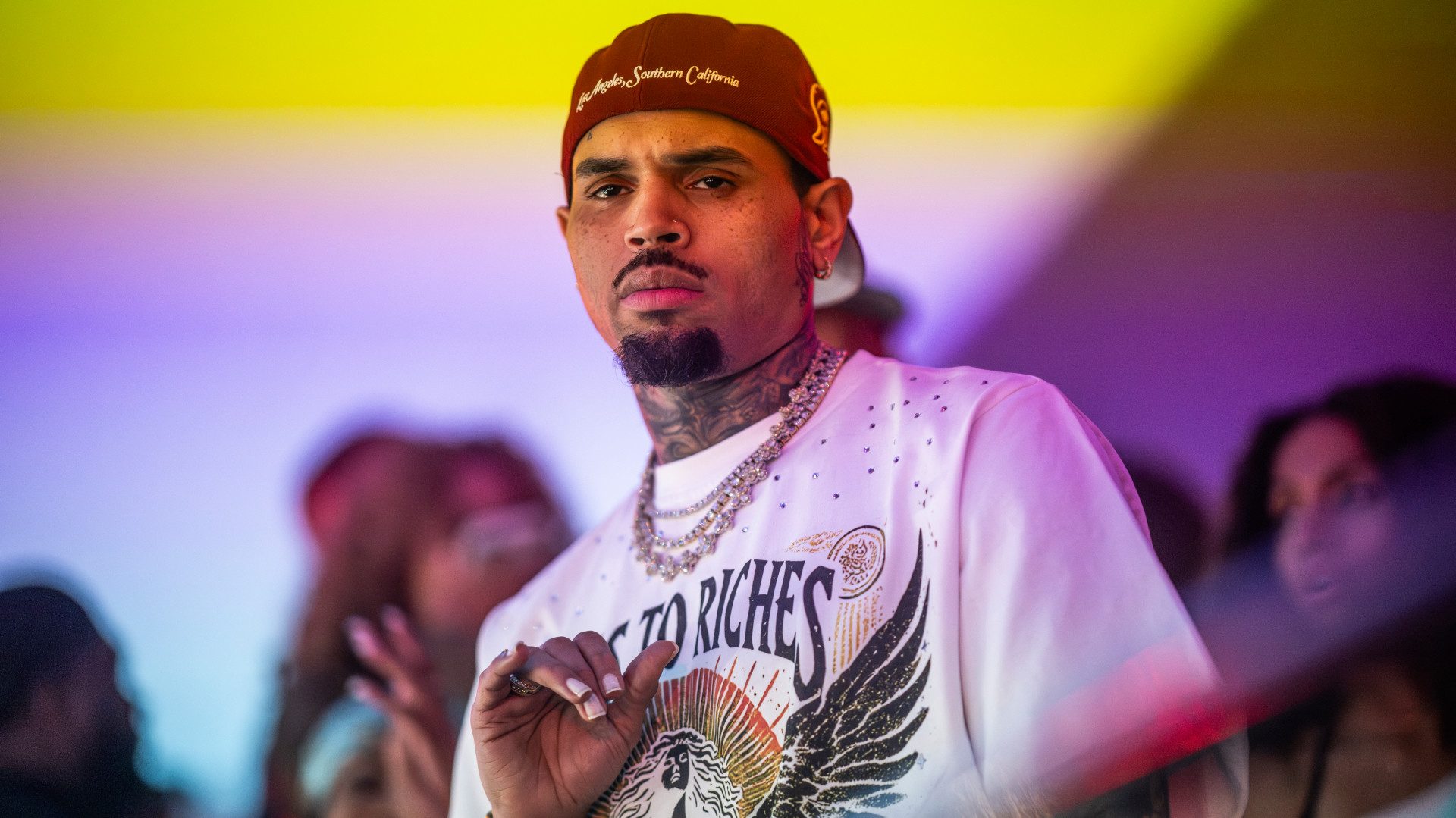 Chris Brown wearing a red cap and white graphic t-shirt, standing in a colorful, blurred background.