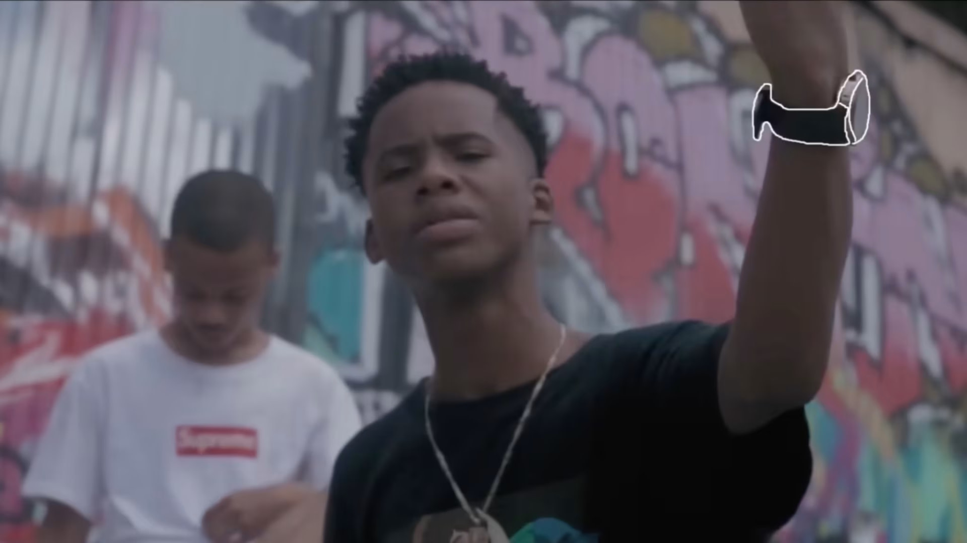 Two young men in casual clothing stand in front of a colorful graffiti wall. One wears a Supreme shirt, and the other gestures with his hand.