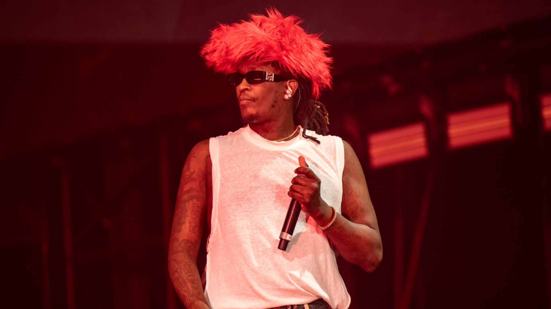 Young Thug on stage wearing a red feathered hat, sunglasses, and a white sleeveless shirt, holding a microphone.