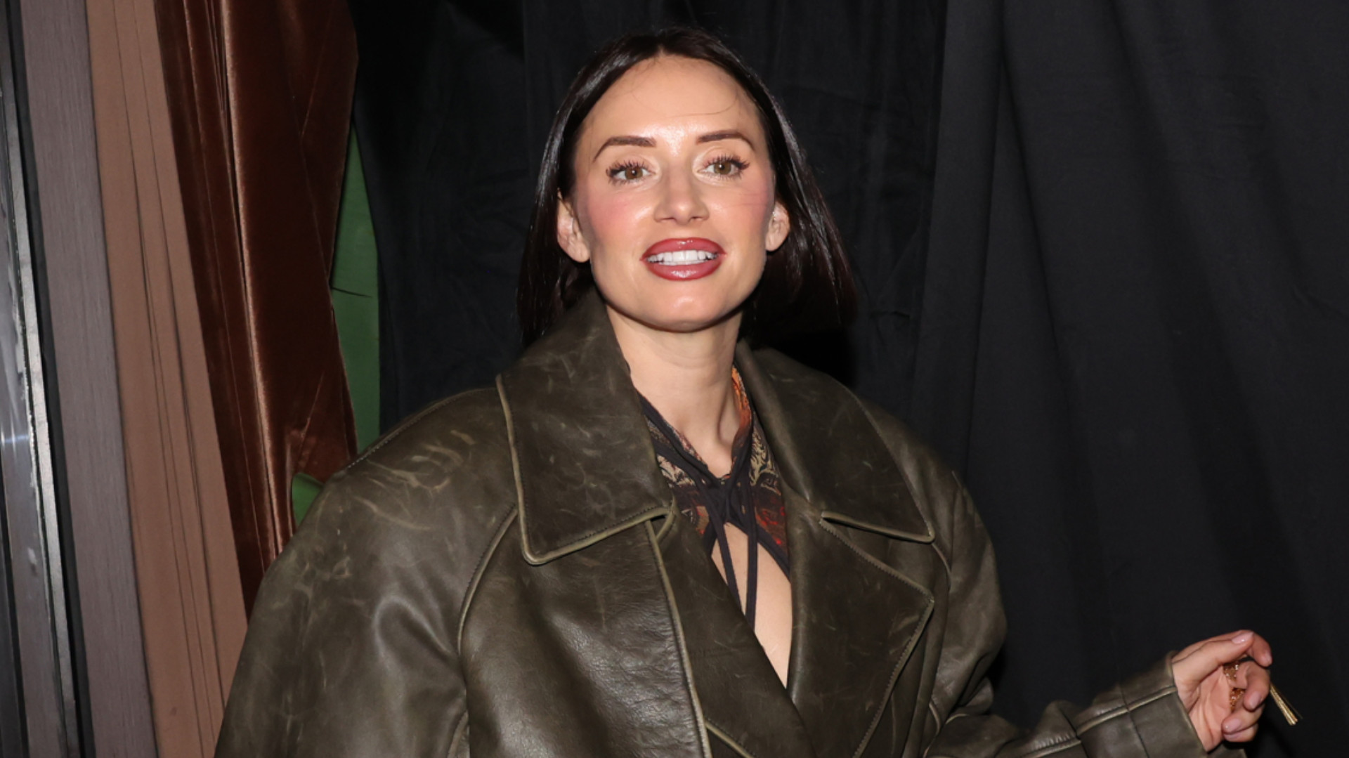 YesJulz with dark hair wearing a large green leather jacket stands in front of a dark background.