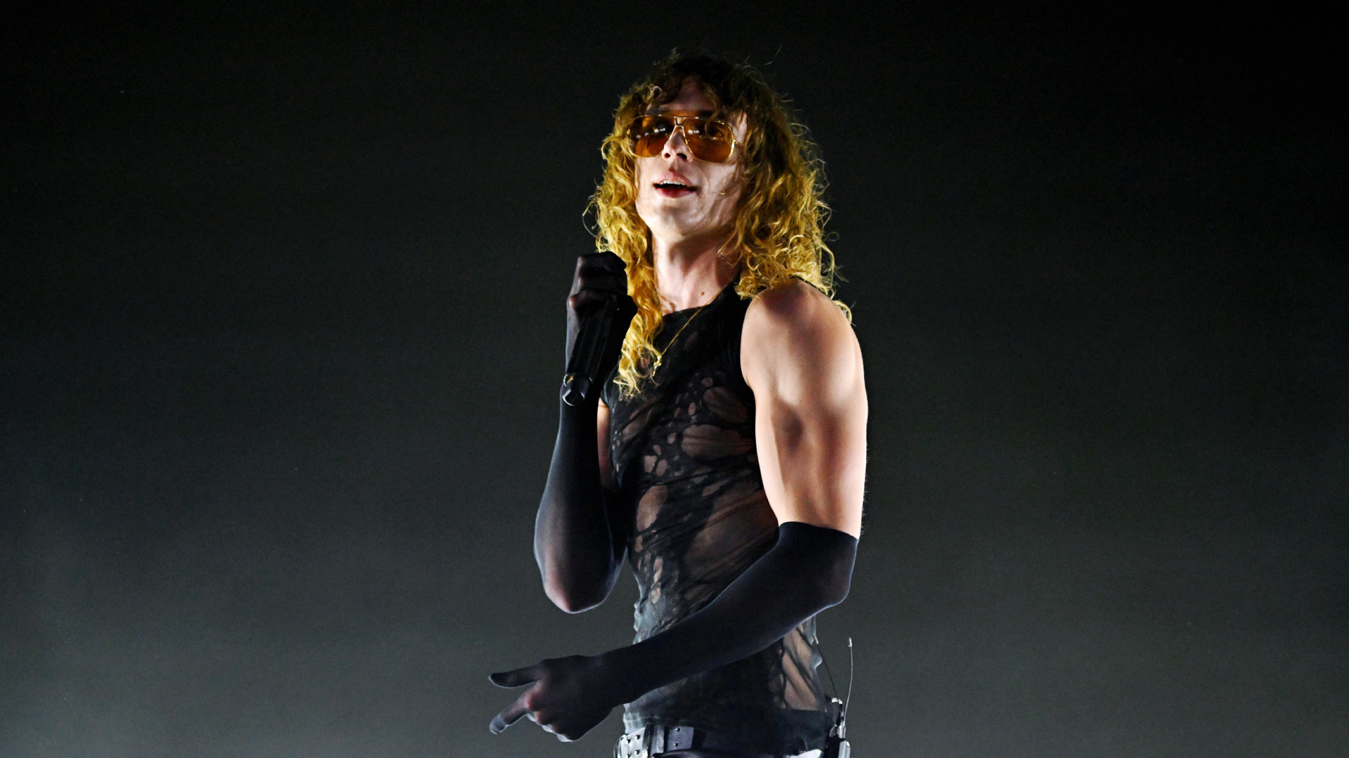 2hollis with curly hair and sunglasses sings on stage, wearing a sheer black top and long gloves.