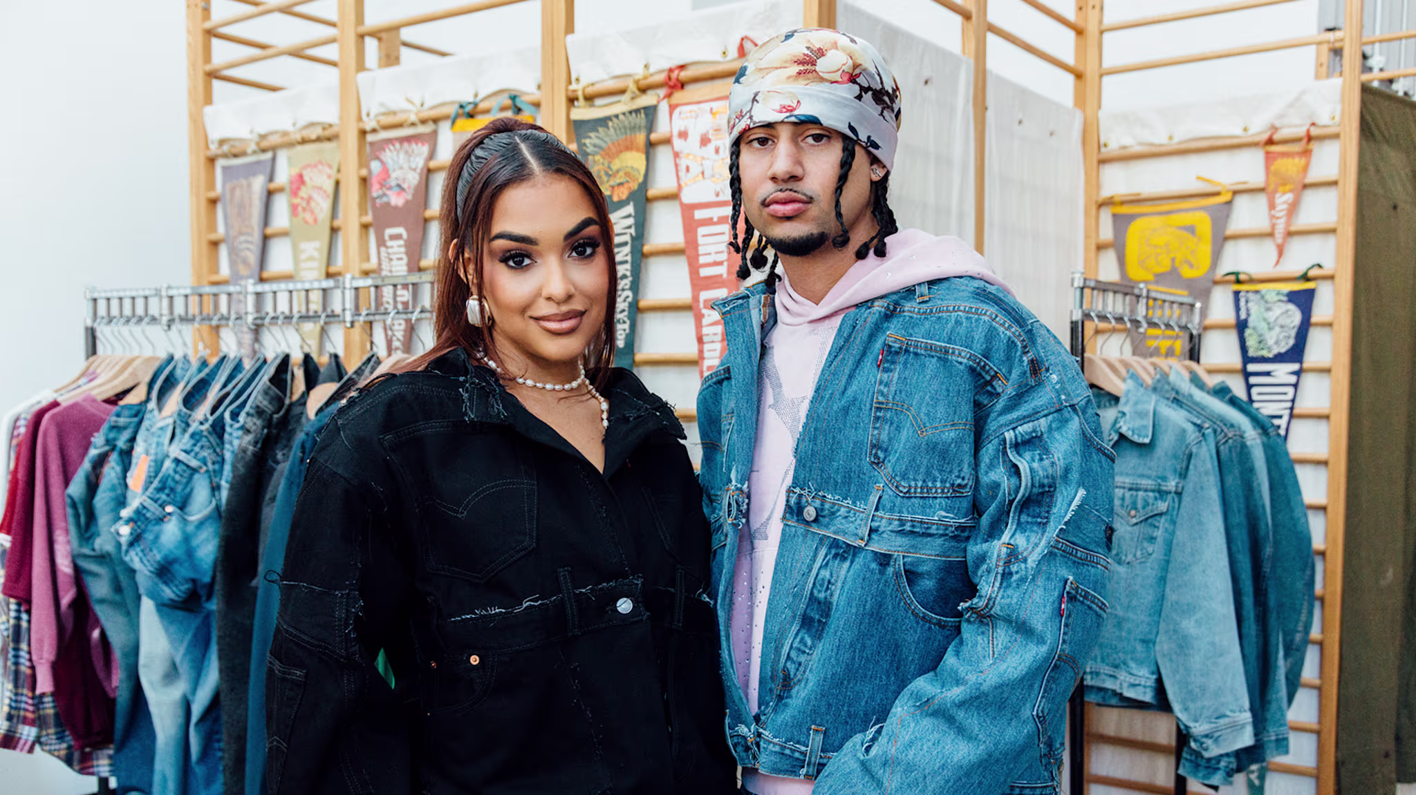 A woman and a man pose in a clothing store. The woman wears a black jacket, and the man wears a denim jacket and a bandana.