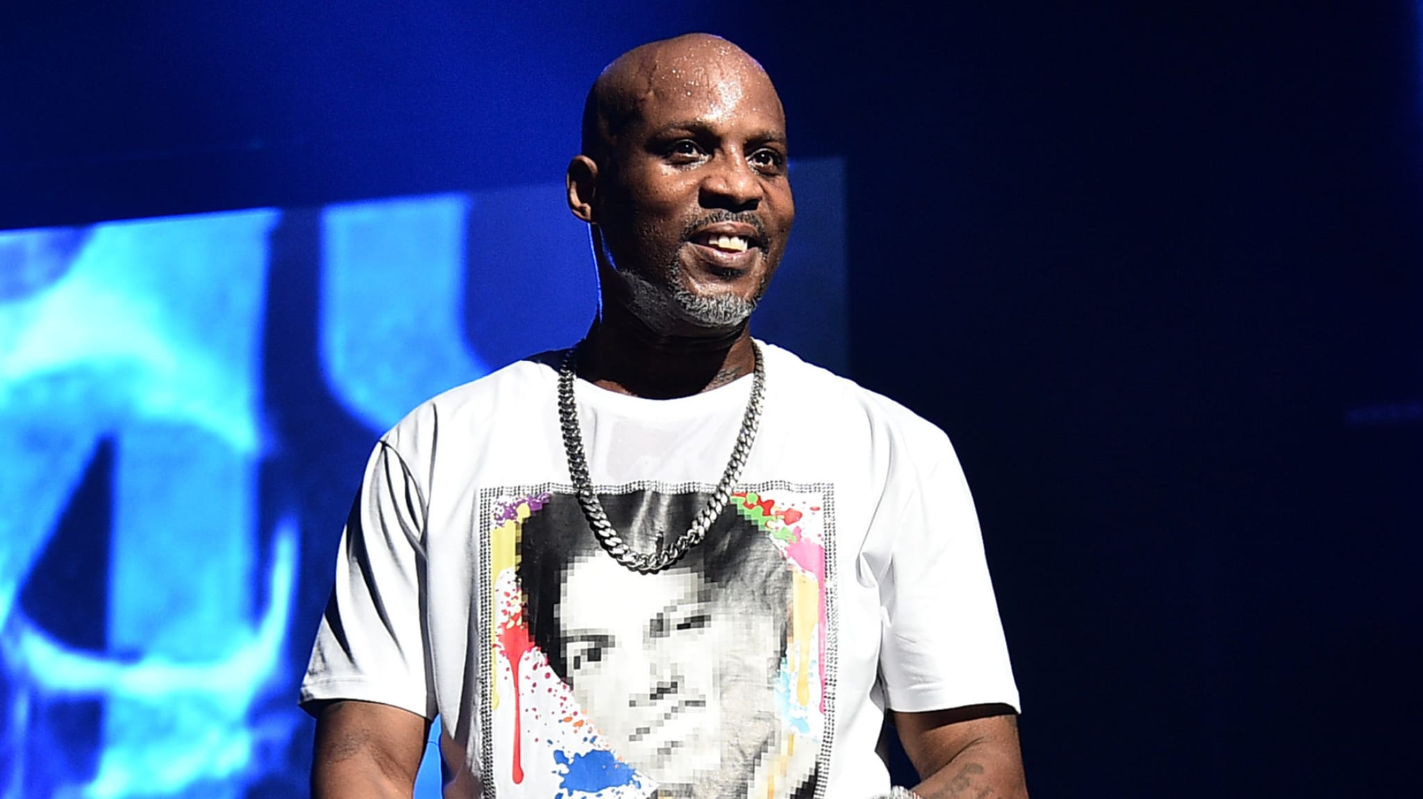 DMX on stage, wearing a graphic t-shirt and chain necklace, with a blue-lit background.