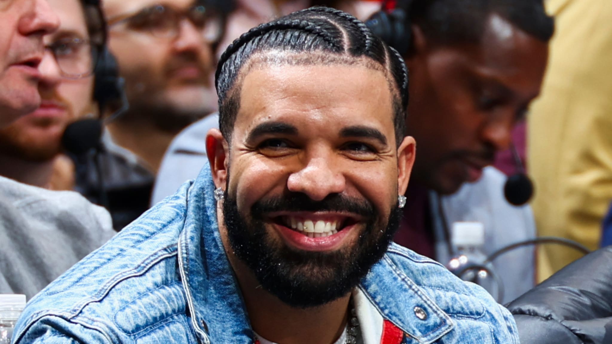 Drake, wearing a denim jacket, smiles while sitting in a crowd.