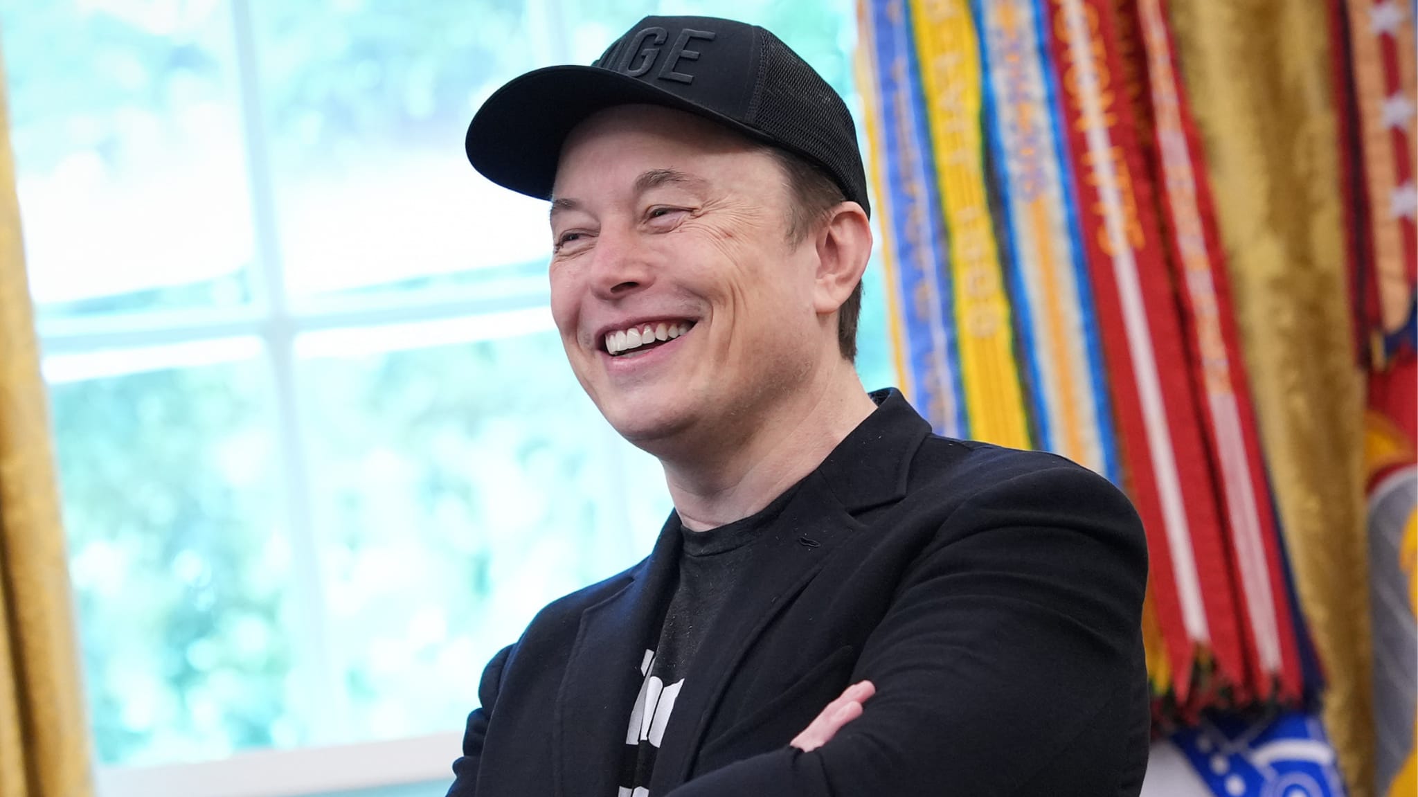 Elon Musk smiling, wearing a black cap and jacket, in a room with colorful decorations in the background.