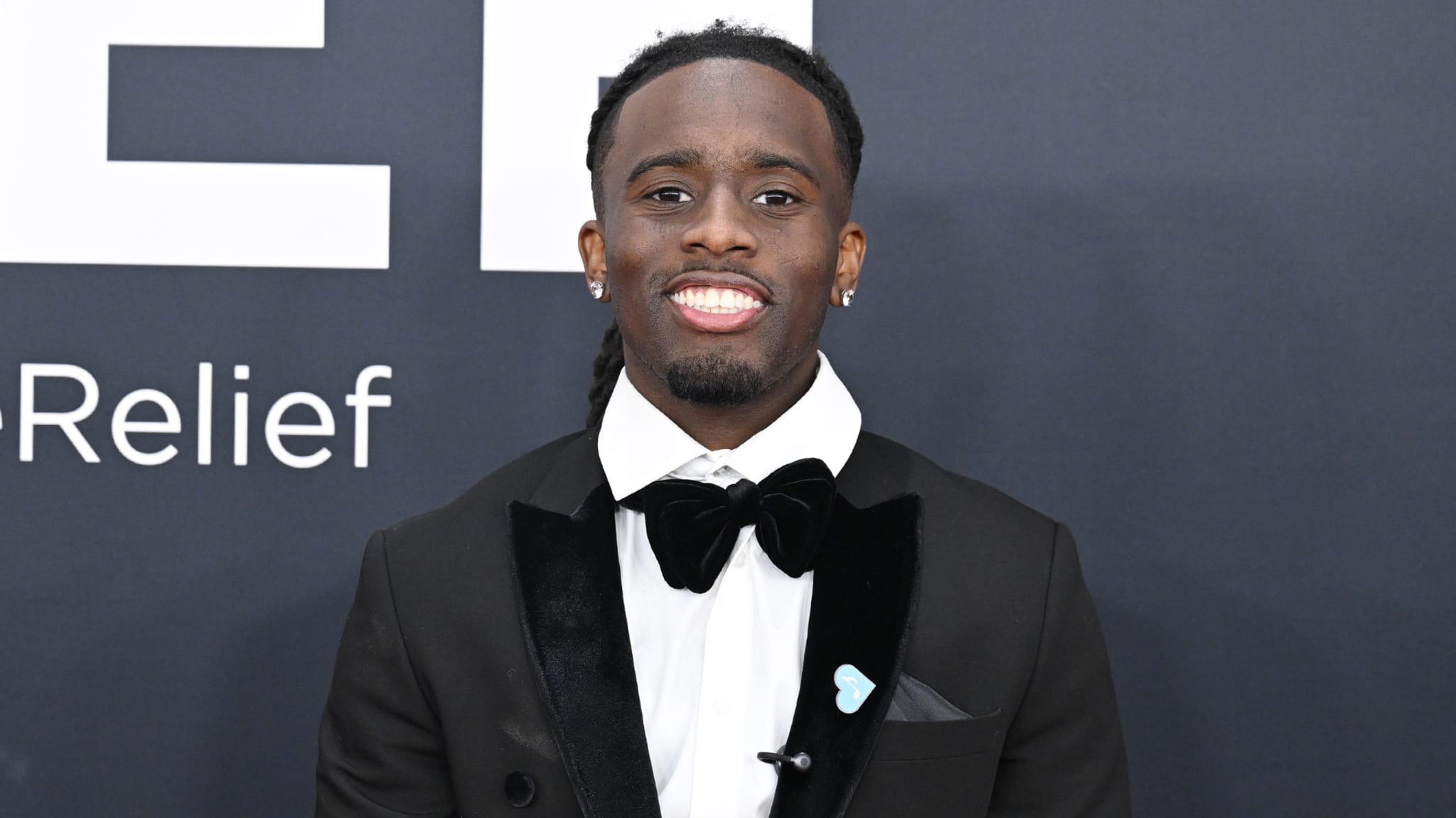 A man in a tuxedo with a bow tie smiles at an event, standing against a dark background with text partially visible.