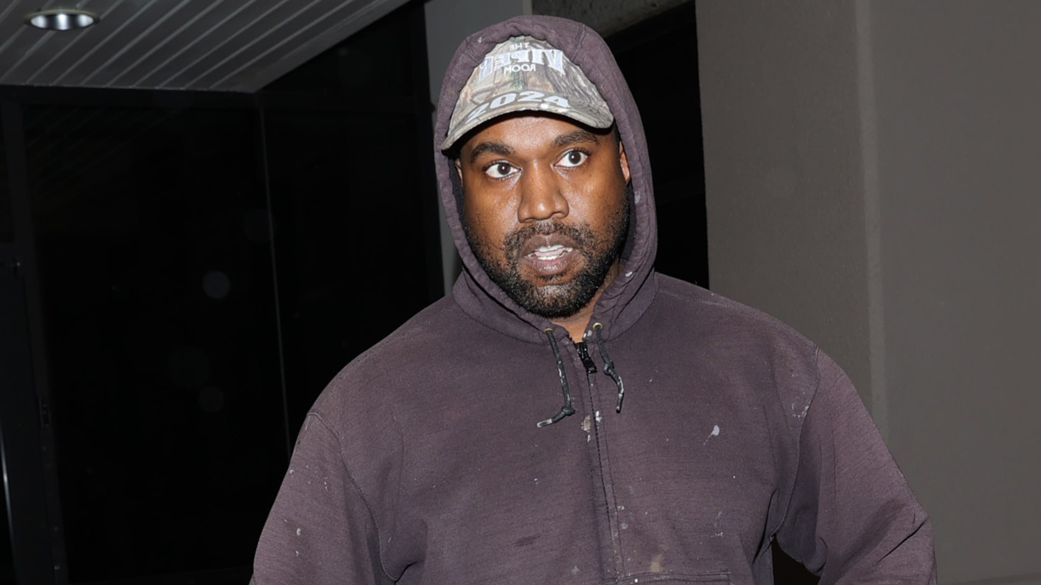 Kanye West wearing a dark hoodie and a cap, looking directly at the camera in an indoor setting.