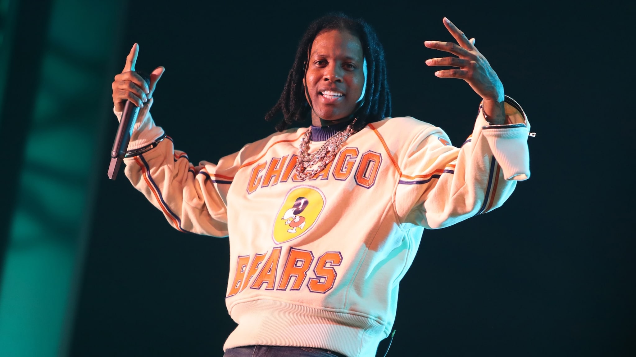 Lil Durk performing on stage, wearing a "Chicago Bears" sweatshirt and holding a microphone, with a backdrop of stage lights.