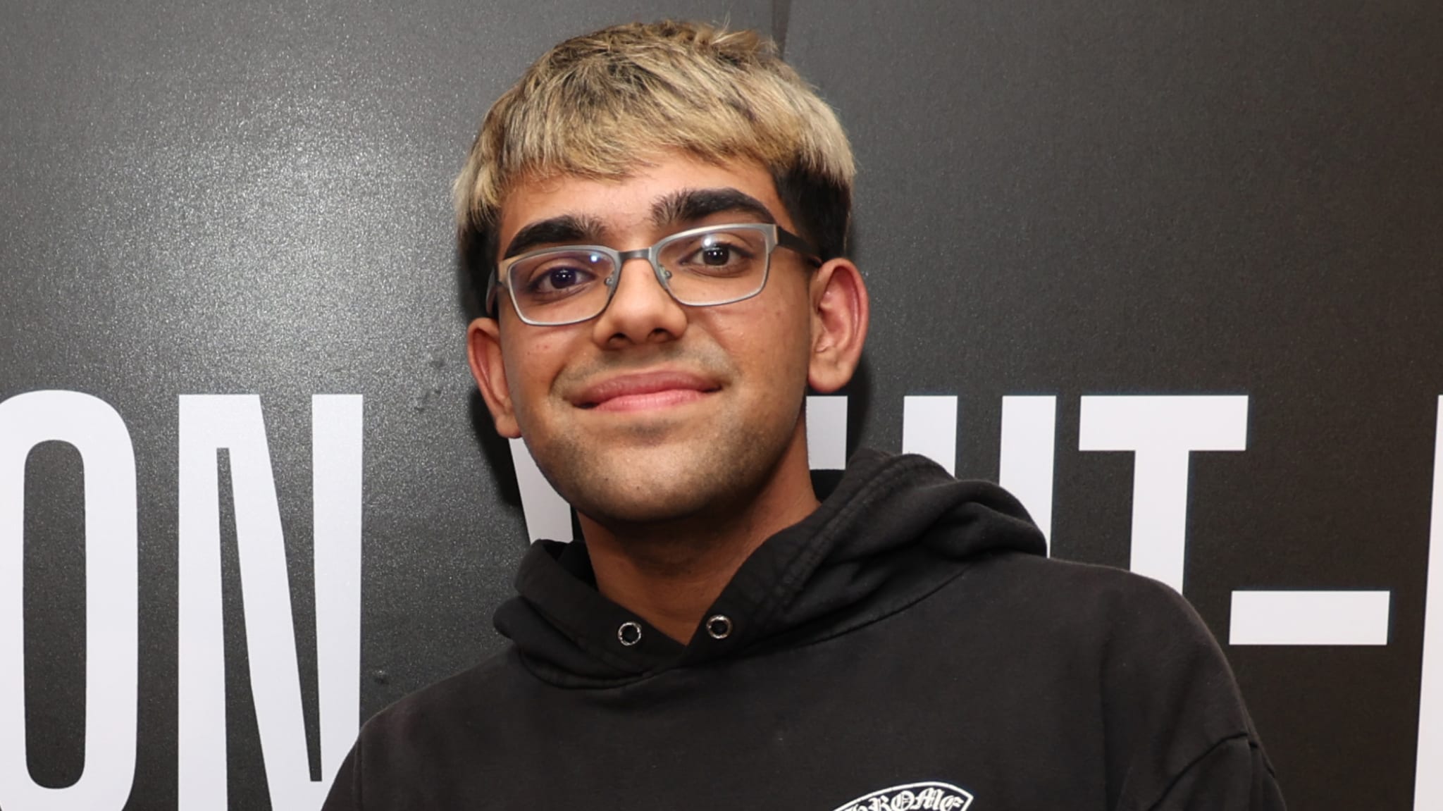 N3on with glasses and short hair stands in front of a black and white background, wearing a black hoodie.