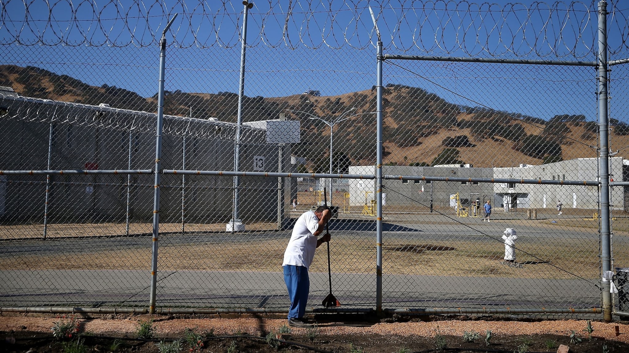 A person in a white uniform leans on a broom near a chain-link fence topped with barbed wire, with prison buildings and hills in the background.