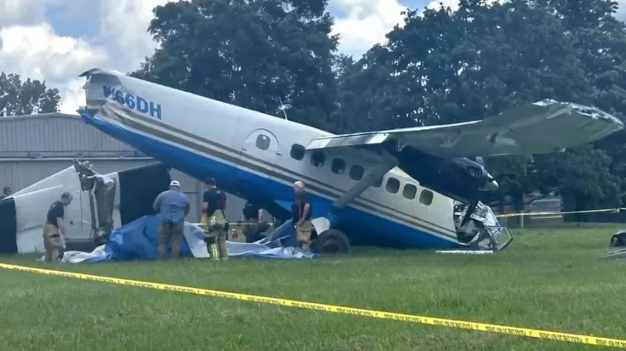 A small airplane with registration N66DH is crashed on a grassy area. Several people in uniforms are inspecting the scene.