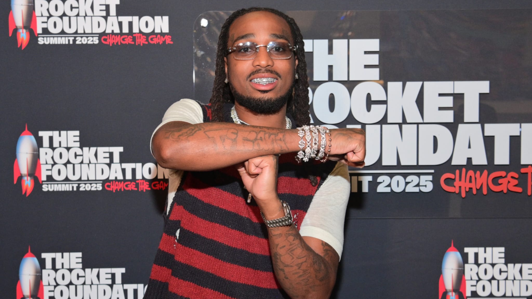 Quavo poses at The Rocket Foundation event, wearing glasses, a striped shirt, and jewelry, in front of a branded backdrop.
