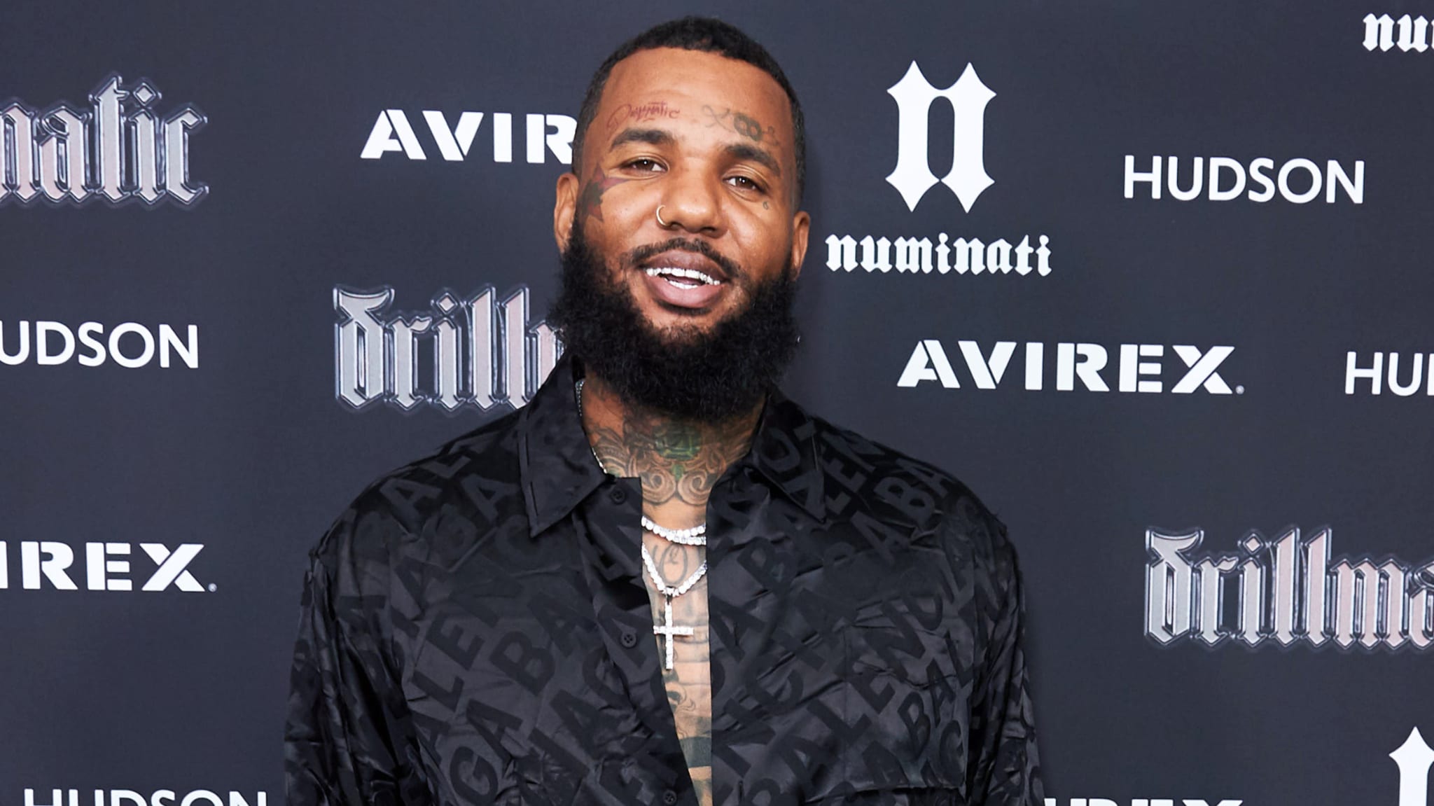 A man with tattoos and a beard is smiling, wearing a patterned black shirt, standing in front of a backdrop with logos and text.