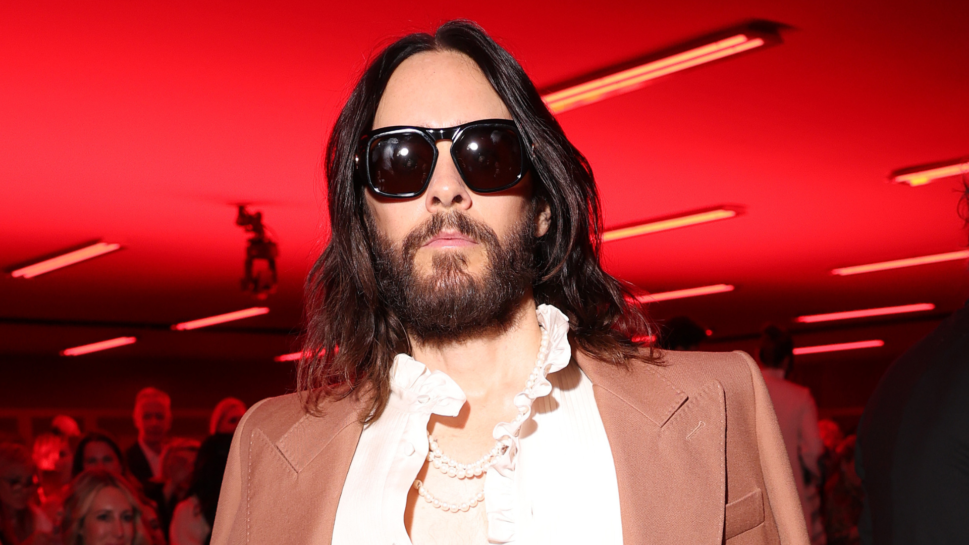 Jared Leto in a stylish tan suit, white shirt, and sunglasses, with a red-lit background.