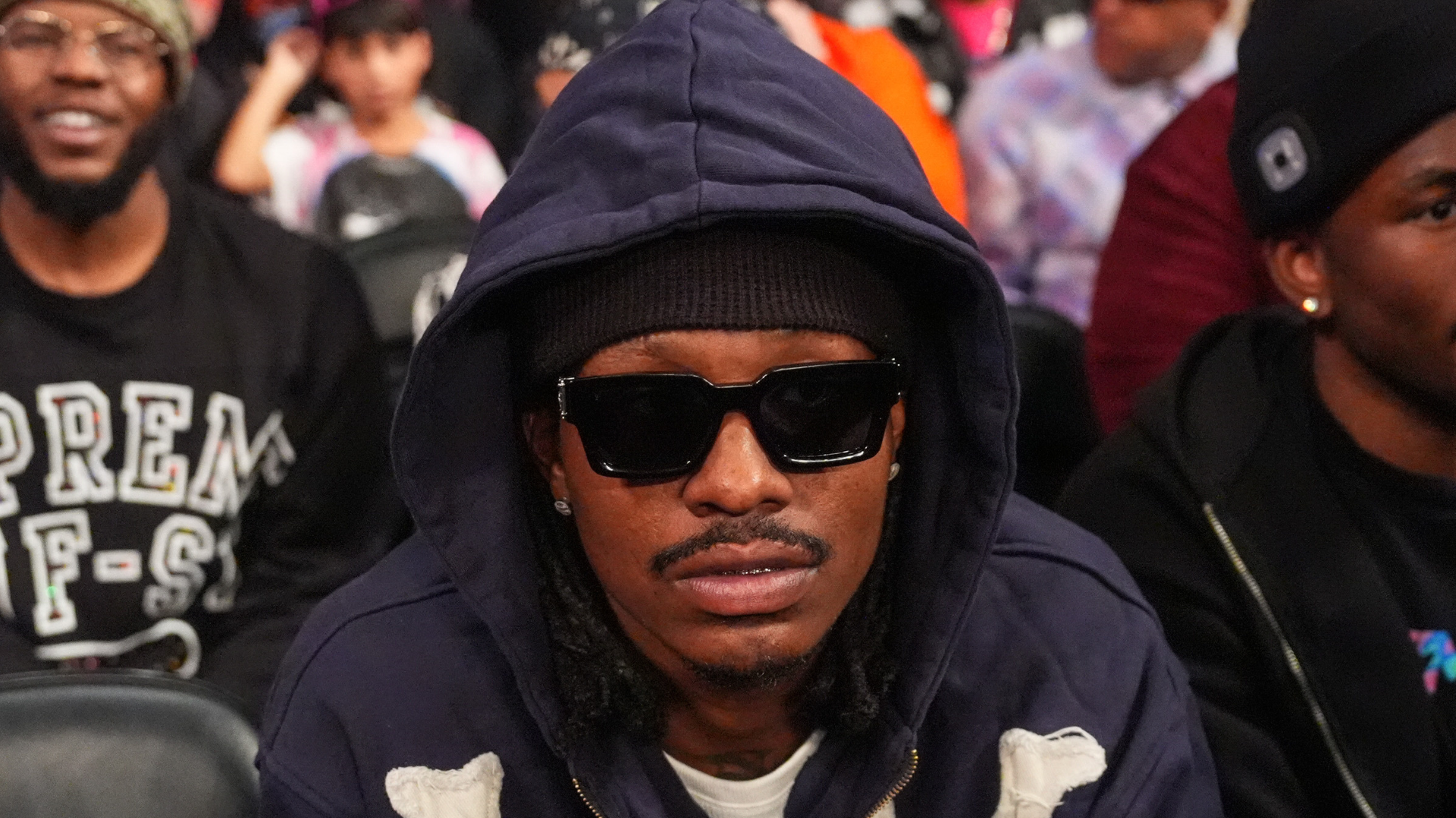 A person wearing sunglasses and a hoodie sits in a crowd, looking towards the camera.