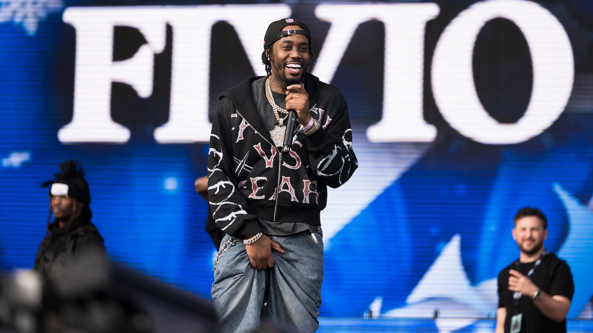 Rapper Fivio Foreign performs on stage, wearing a black jacket and jeans, with a large screen displaying "FIVIO" behind him.