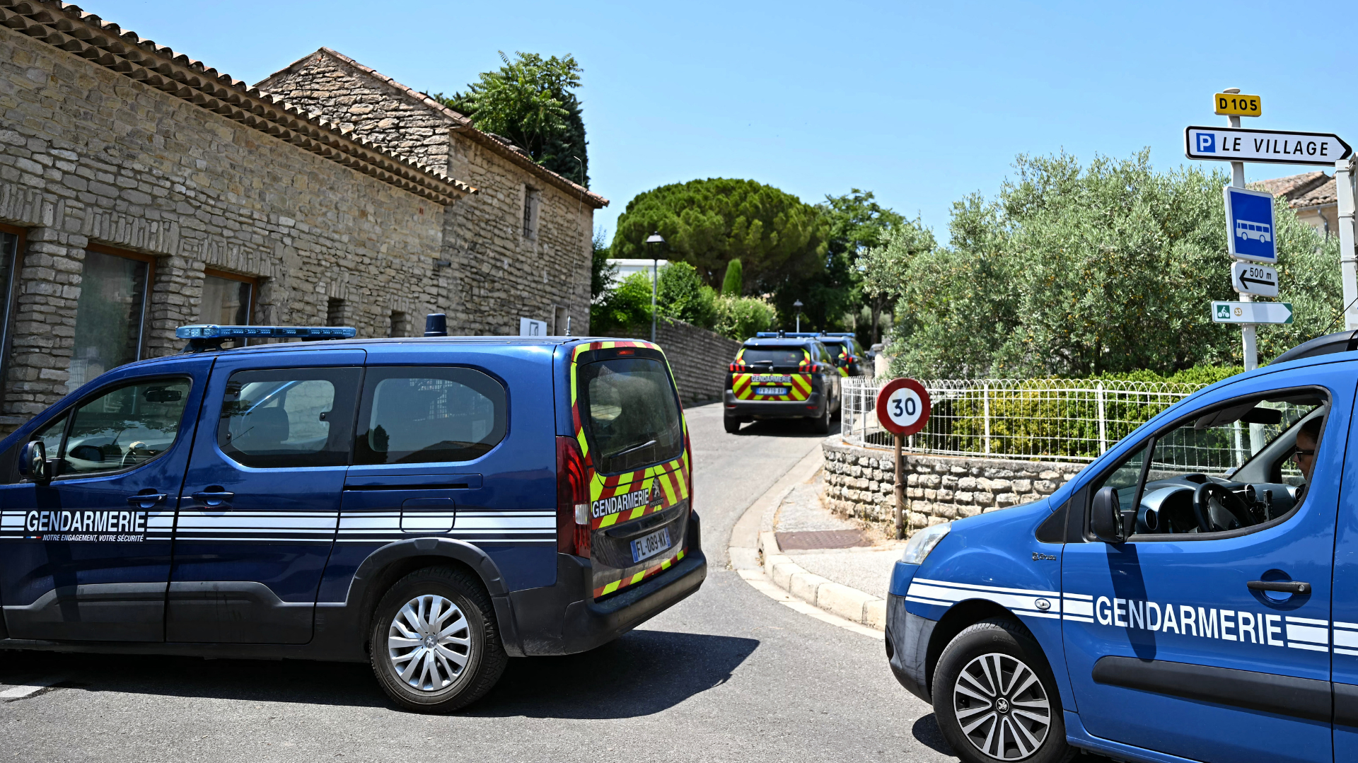 Gendarmerie vehicles are parked on a narrow street in a village setting, with stone buildings and greenery in the background.