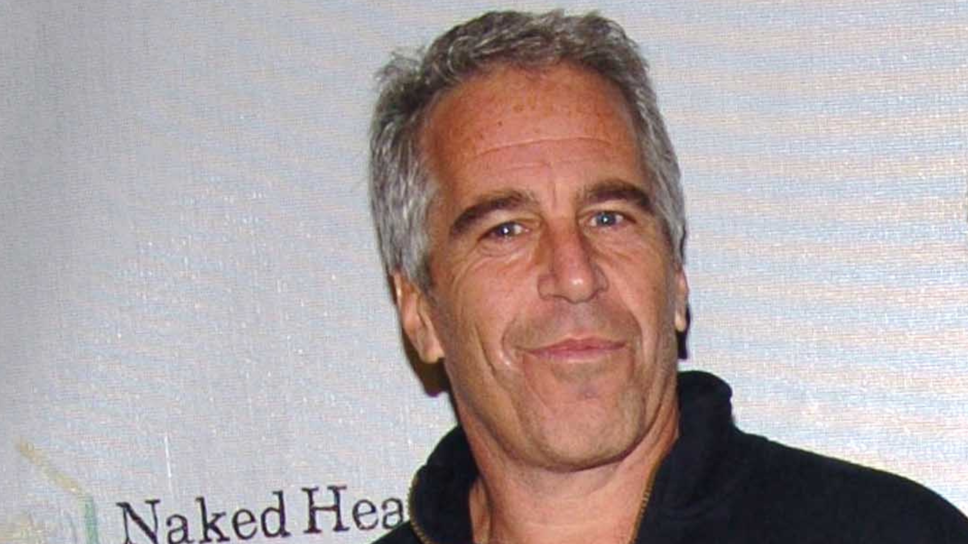 A man with gray hair and a black jacket stands in front of a backdrop with partial text visible.