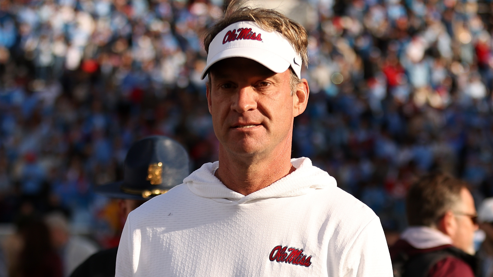Lane Kiffin wearing an "Ole Miss" visor and hoodie stands in a stadium, with a crowd in the background.
