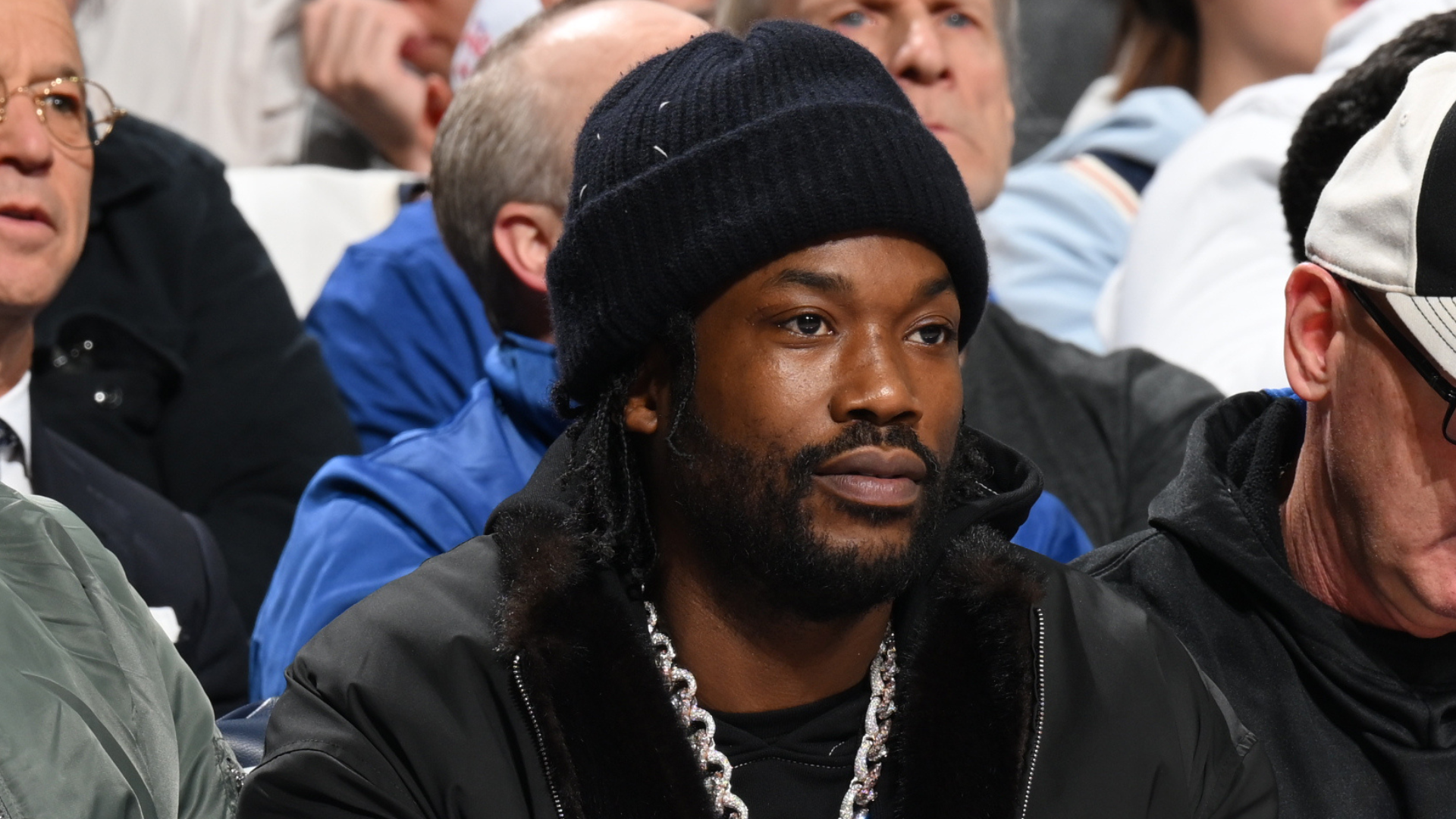 Meek Mill wearing a black beanie and chain necklace sits among a crowd at a sports event.