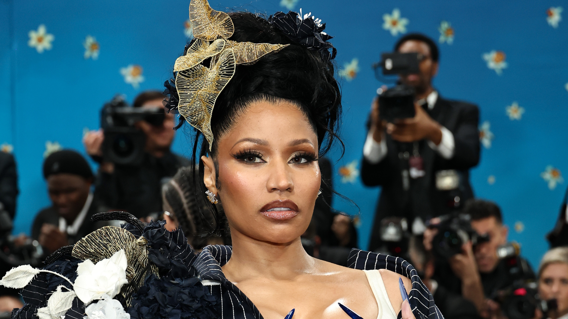 Nicki Minaj at a formal event, wearing an elaborate headdress and dark outfit, with photographers in the background.