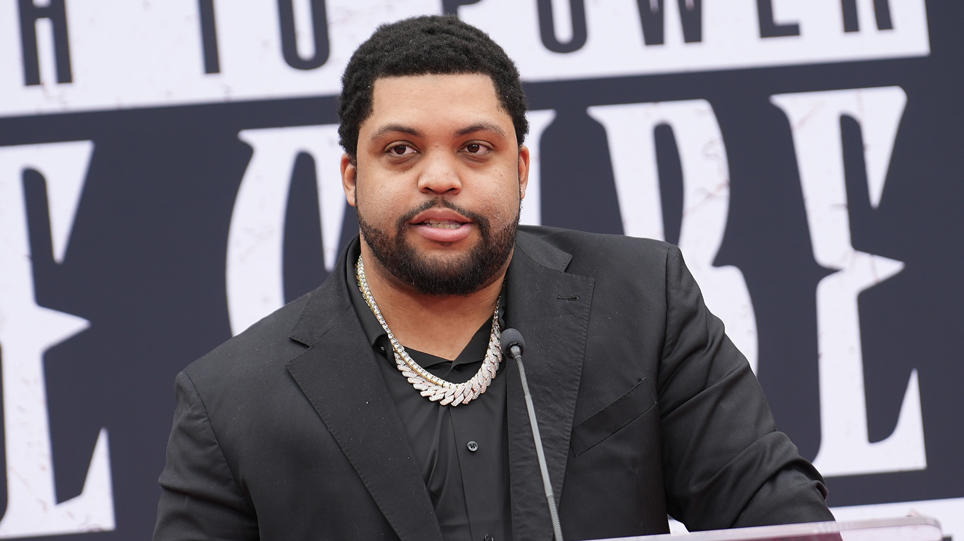 A man with a beard and short hair, wearing a black suit and a necklace, speaks at a podium with a microphone.