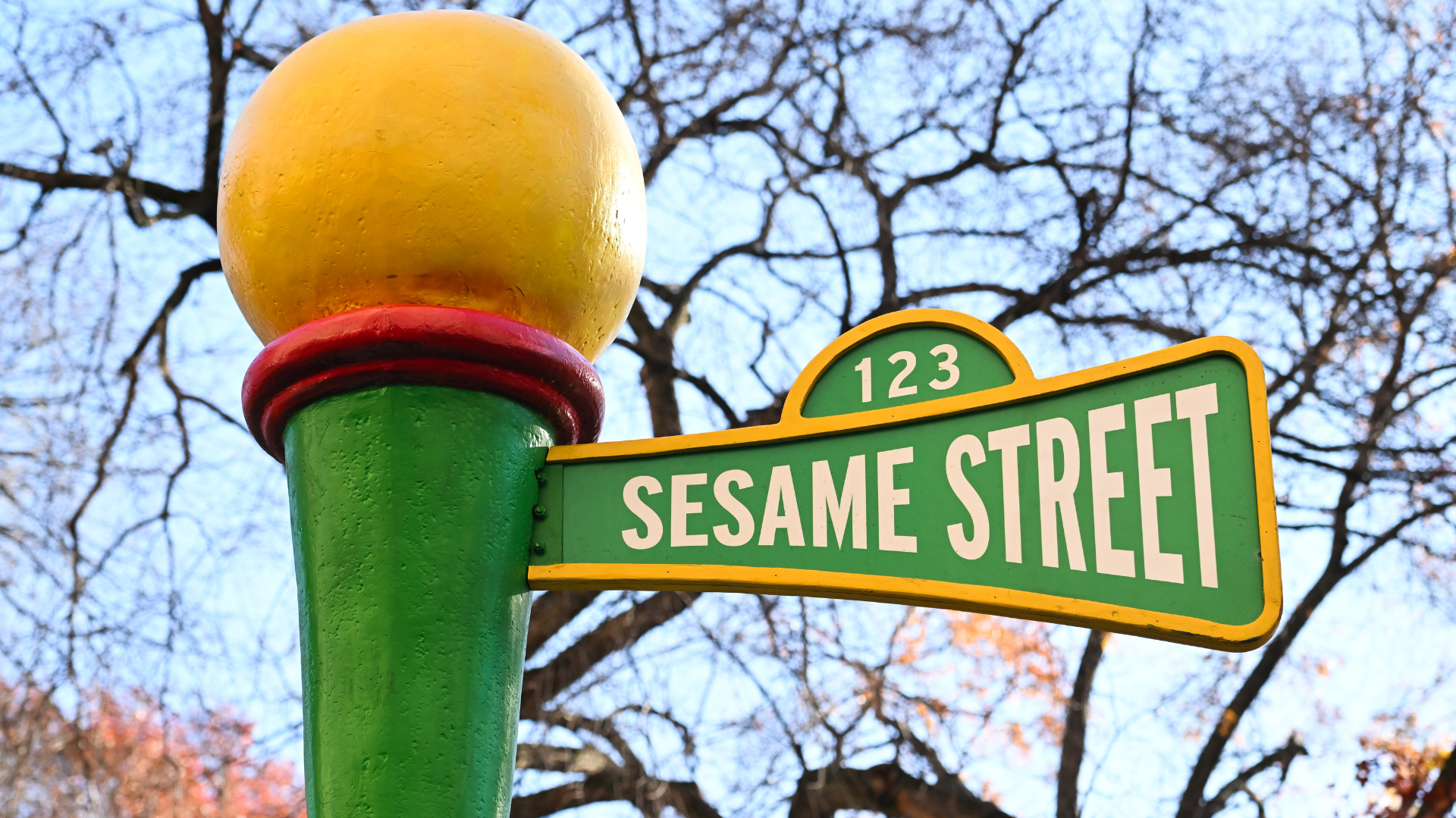 A green street sign with "123 Sesame Street" in white letters, set against a backdrop of bare tree branches.