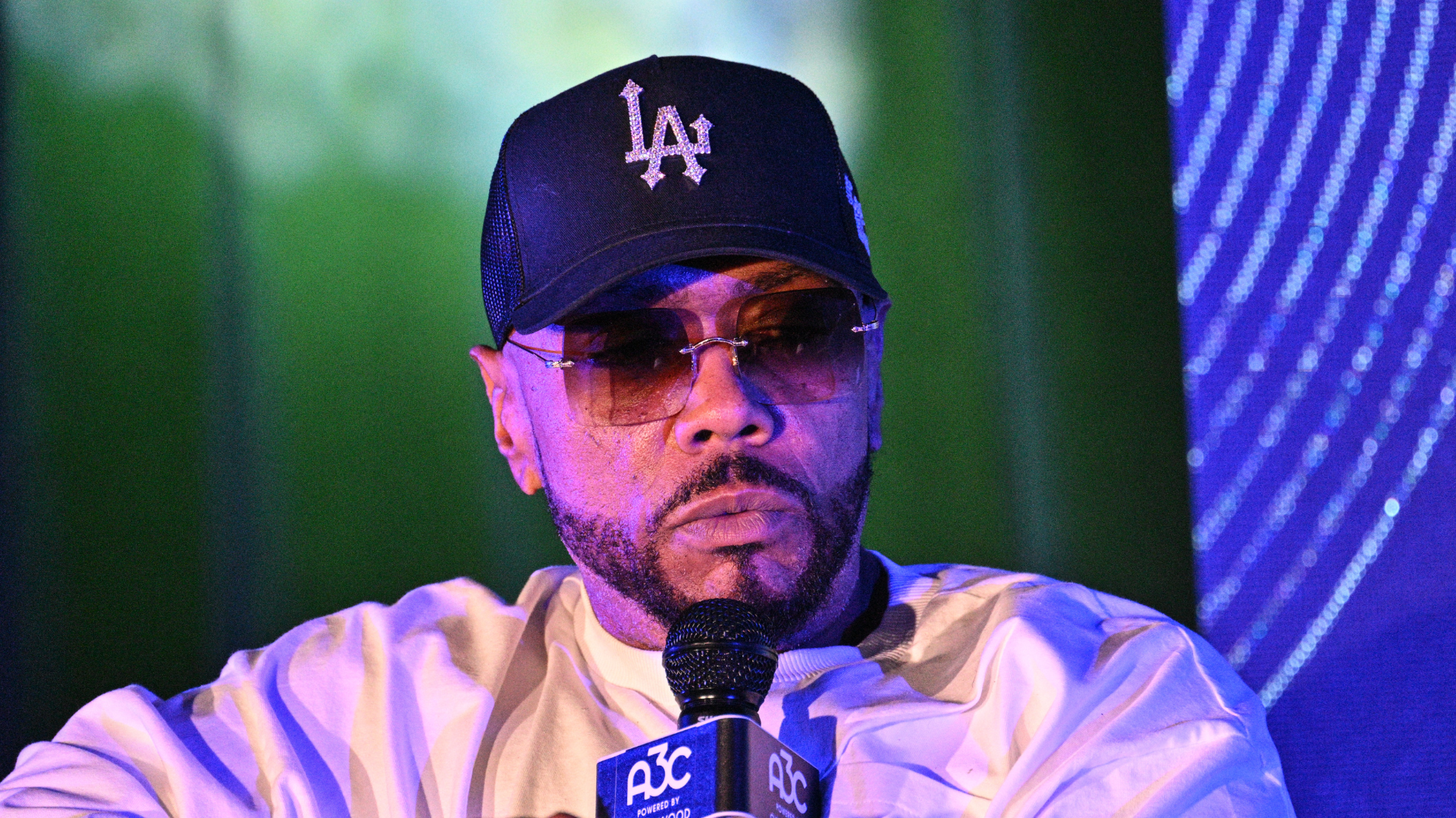 Man in a cap and sunglasses speaking into a microphone at an event.