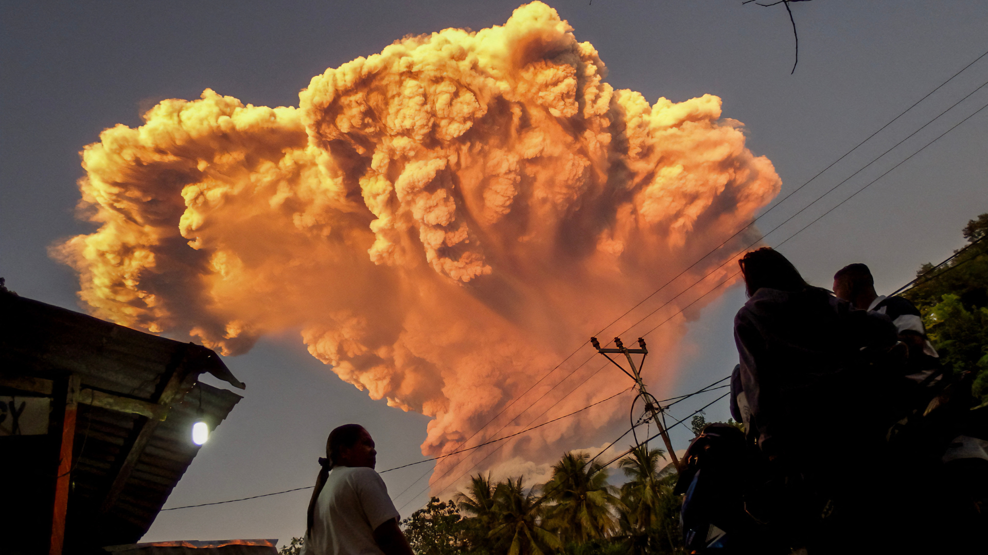 A large volcanic eruption with a massive plume of smoke and ash in the sky, silhouetted people watching, and palm trees below.