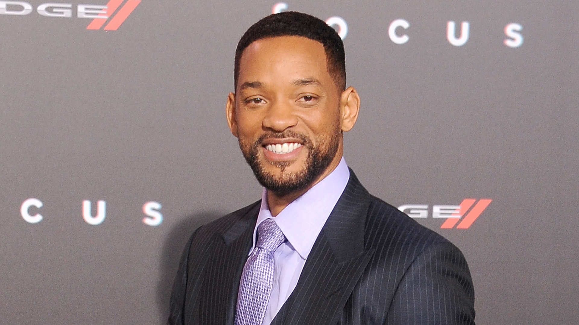 Will Smith smiling at an event, wearing a dark suit and light purple tie, with a "Focus" backdrop behind him.