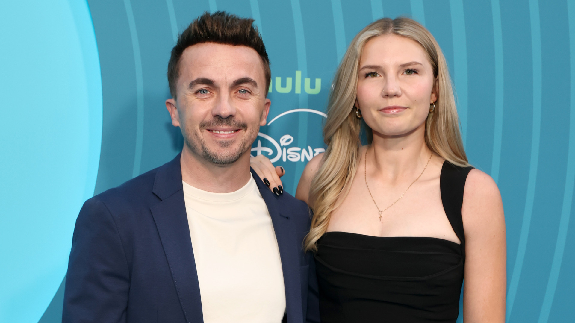 Frankie Muniz in a blue blazer and wife Paige Price in a black dress pose together against a blue backdrop.