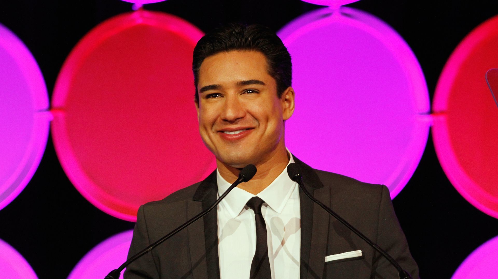 Mario Lopez, in a suit, smiles at a podium with colorful circular lights in the background.