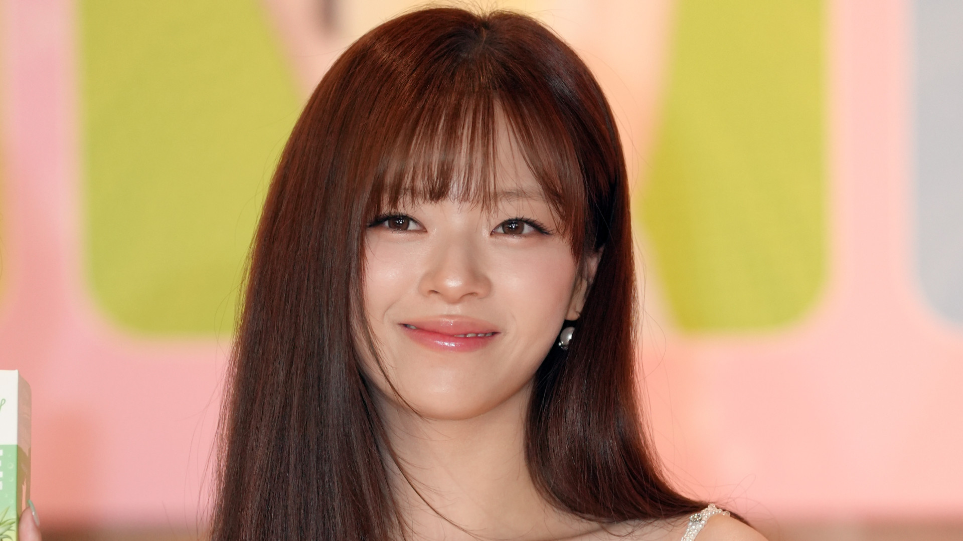 TWICE's Jeongyeon with long brown hair and bangs, smiling in front of a colorful background.
