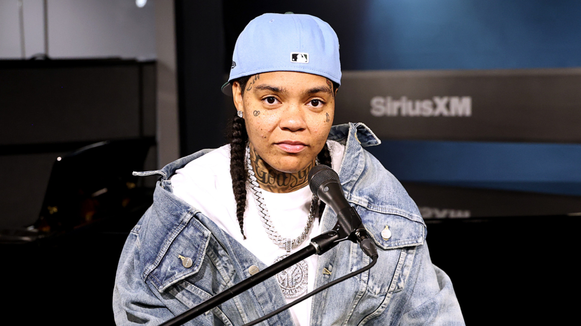 Young M.A, wearing a blue cap and denim jacket, sits at a microphone in a recording studio.